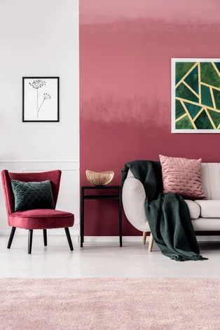 Illustration - Ihr Experte für schönes Wohnen - Feminines Wohnzimmerinterieur mit bordeauxrotem Sessel und dunkelgrüner Decke auf einem beigen Sofa, das an einer Ombre-Wand steht